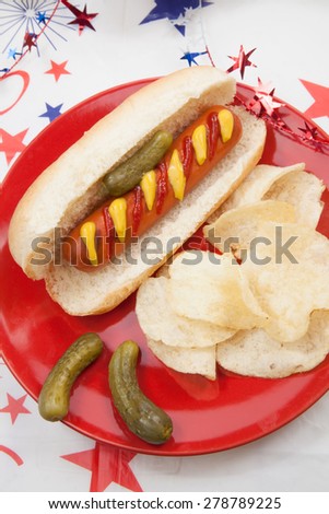 Hot dog, chips, and pickled cucumbers. Angel food fruit cakes and muffins on 4th of July in patriotic theme in background.