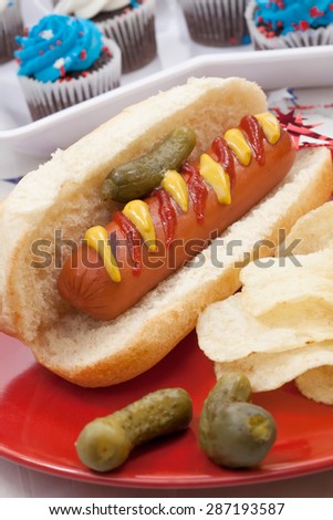 Hot dog, chips, and pickled cucumbers. Angel food fruit cakes and muffins on 4th of July in patriotic theme in background.