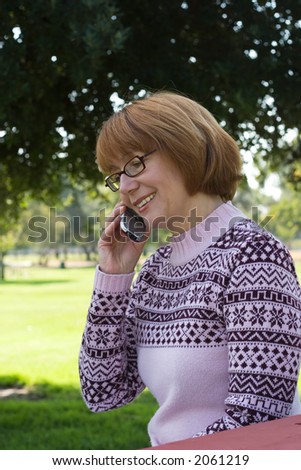 Smiling woman is talking on the cell phone on good day outside in the park with trees in the background