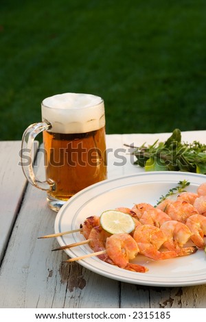 Grilled shrimps on wood sticks served on outside table with lime and mug of pale ale