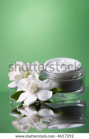 Closeup of container of opened moisturizing face cream and blooming fragrant white gardenias on melted icecubes over green toned background