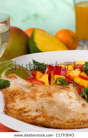 Closeup of fried skate (ray) with mango, cherry tomato and red onion salsa. Fresh fruits, glass of white wine and glass of juice.