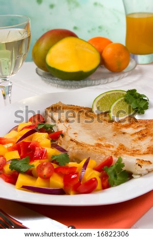 Closeup of fried skate (ray) with mango, cherry tomato and red onion salsa. Fresh fruits, glass of white wine and glass of juice.
