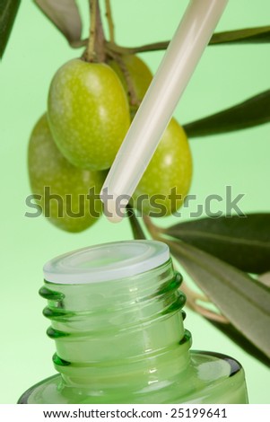 Closeup of dropper with drop of face cream and branch of green olives (out of focus) over green background