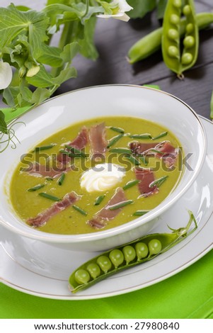 Bowl of pea soup with prosciutto garnished with chives. Sprig of flowering pea and cracked pea pods.