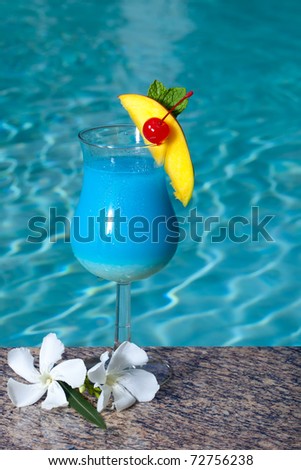 Glass of Blue Hawaiian cocktail on swimming pool side garnished with mango wedge and maraschino cherry