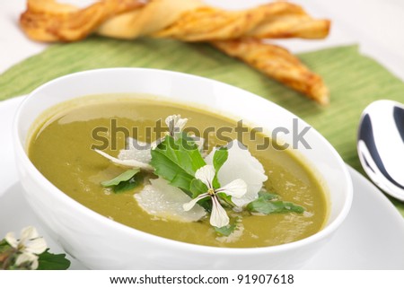 Bowl of pea soup garnished with rocket / arugula leaves and flowers, and shaved Parmesan cheese. Cheese sticks in background.