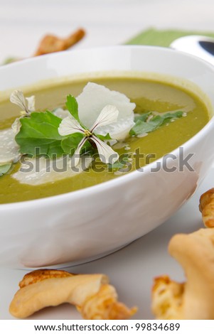 Bowl of pea soup garnished with rocket / arugula leaves and flowers, and shaved Parmesan cheese. Cheese sticks in background.