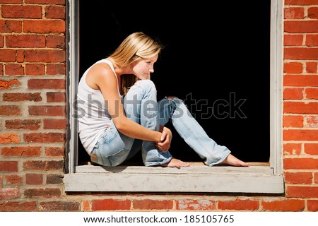 Beautiful young woman sits on window ledge of abandoned brick building.