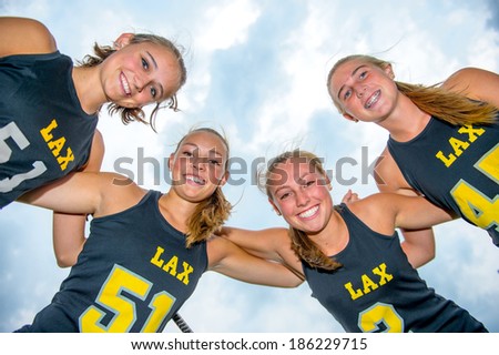 Perspective looking up with team of girls lacrosse players huddled close together, arms on each others shoulders, smiling and looking down at the viewer.