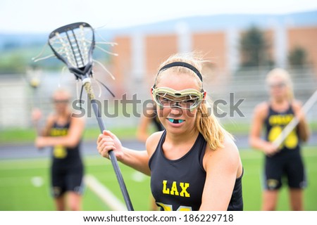 Aggressive women\'s lacrosse player with teammates in background. She is wearing full protective gear and holding her lacrosse stick.