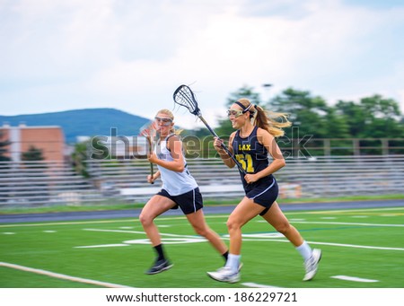 Panning blur featured as two female lacrosse players race down the field. High degree of motion conveyed. Some motion blur on athletes, particular in extremities, focus on face of closest player.