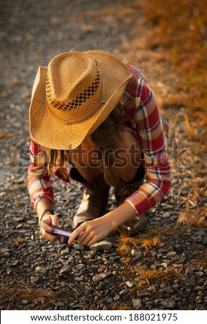 Young woman wearing straw hat, red plaid shirt, and cowgirl boots using a pocket point and shoot camera to capture a detail on the farm.