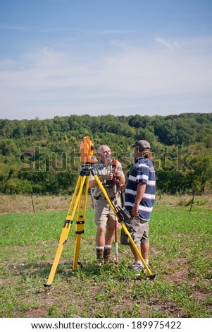 Two man survey crew working outdoors on a sunny day. They converse near an electronic distance meter and tripod.