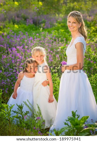 Bride with her flower girls in lush green and flower filled field.