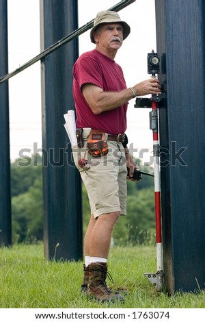 One worker of a two man survey crew holds a prism at the foundation to record its position. He has rolled up blueprints in back pocket and various surveying tools on his utility belt.