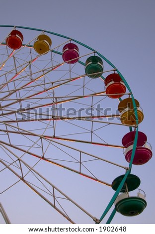 Colorful cars on a ferris wheel seen against a darkened sky, but before nightfall. Cars are empty and colored lights glow on the spokes of the wheel.