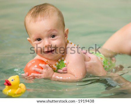 Six month old baby girl at her first swimming lesson. Smiling and looking at the viewer, also holding an orange floating toy.