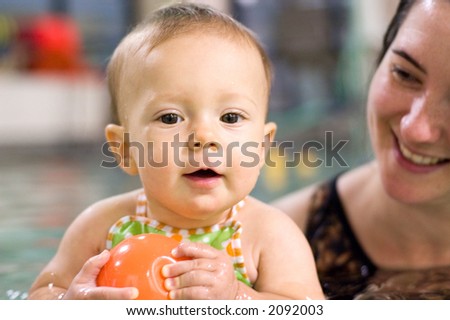 Baby girl holding an orange ball in an indoor swimming pool. Her mother is behind her holding and supervising the lesson.