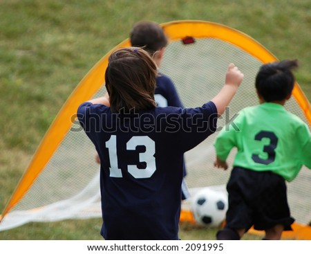 Young soccer player celebrates as teammate scores a goal. Strong DOF focus on closest player with ball and goal seen as smooth backdrop.
