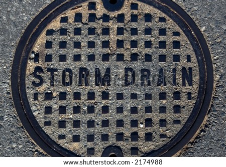 Manhole cover implanted in a paved asphalt road. Metal grid and large lettering stamped with words Storm Drain.