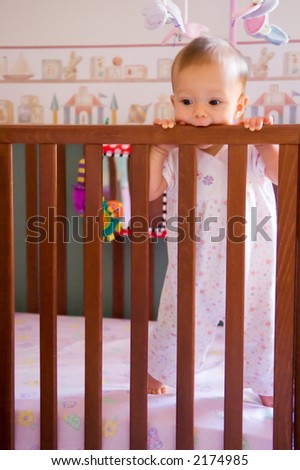 Baby less than one year old standing in her crib and chewing on top rail, cutting teeth.
