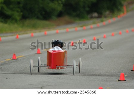 Red/Orange soap box race car speeds down the track. Shallow depth of focus on car and driver while forest background becomes a blur.