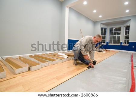 Man using a rubber mallet and tapping block to install the next row of laminate flooring. Do-it-yourself and renovation themes enhanced by uncovered wall outlets and power tools in the background.