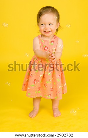 Baby girl in Spring dress smiles and claps while she looks to the left as soap bubbles float all around her. Yellow Background.