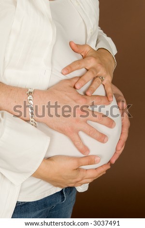 Expectant young mother in third trimester cradles her belly with dad's hands wrapped around too. Warm brown background.