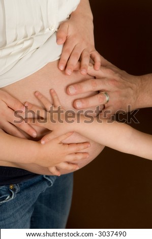 Mother in third trimester with children\'s and husbands hands touching her belly. DOF focus on hands and top of belly with blurring on the underside of belly and jeans.