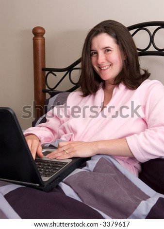 Young woman working from the comfort of her own bed. Wearing pink terry cloth robe while checking e-mail on a laptop.