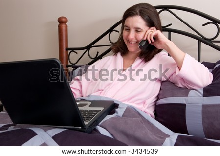 Lucky woman enjoys telecommuting from the comfort of her own bed. She\'s even wearing a terry cloth robe as she talks on the phone and accesses her laptop.