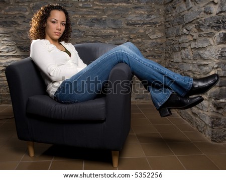 Beautiful young woman in casual clothing, relaxing in a chair with a stone wall interior in the background.