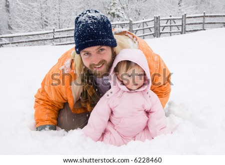 Proud father with one year old baby girl playing in her first snow. He is smiling. She looks timid. Snow still falling. Father wearing wool winter cap and a beard. Girl in pink snowsuit.