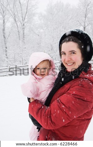 Beautiful new mother and baby girl out for her first time in the snow. Snow still falling. Mother in red coat. Baby in pink snowsuit with hand covers. Picturesque snow covered woods in background.