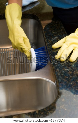 Maid wearing latex gloves and using a large brush to clean a stainless steel kitchen sink in a granite counter top.