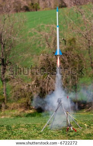Large model rocket blasts off the pad with a long flame emitting from its thruster and plentiful smoke.
