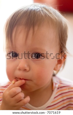 Close-up of a toddler girl in mid-snack. She has a piece of oat cereal on her finger tips and on her mouth.  Shallow f/1.8 DOF focus gives soft quality to the image.