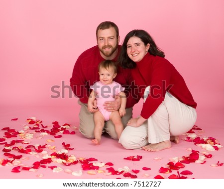New family together for a Valentines Day portrait. Baby in pink and parents in red, against a pink background, with real rose petals.