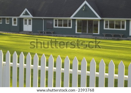 Lawn bowling clubhouse. Focus on white picket fence with green lawn and clubhouse in background.