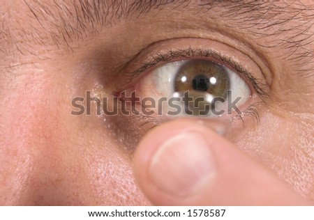 Closeup view of a man\'s brown eye while inserting a corrective contact lens on a finger.  Focus is on the eye and the finger and contact lens are out of focus.