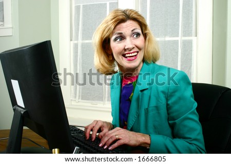Beautiful middle aged executive business woman laughing with off-stage co-workers while typing on her computer in the office.