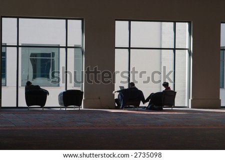 Partial silhouette of three people relaxing, reading, and listening to music in modern style comfortable chairs near a large window in a convention center.
