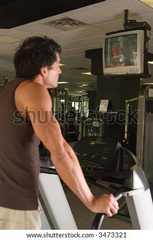 Man working out on a treadmill in a fitness center and watching television while he exercises.  The man is out of focus.  The television he is watching is in focus.