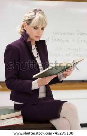 Woman teacher or instructor in a college, university, high school, middle school, elementary classroom sitting on her desk holding a book and reading.