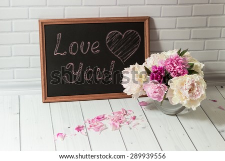 Beautiful bouquet of pink and white peonies in the glass vase with petals and black chalkboard with inscription Love You on the light grey brick and wood background.