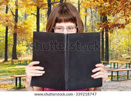 girl with glasses reads big book with blank cover and yellow autumn forest on background
