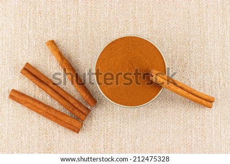 Ground cinnamon powder in porcelain bowl and cinnamon sticks on rustic table cloth, seen from above