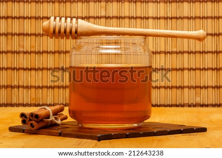 Honey in glass jar with wooden dipper and cinnamon bars on wooden tabletop and mat, with straw mat backdrop